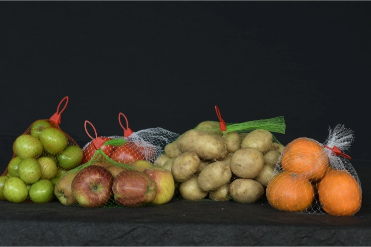 Fresh fruits and vegetables packed in mesh net bags, including apples, potatoes, oranges, and jujubes, displayed against a black background.